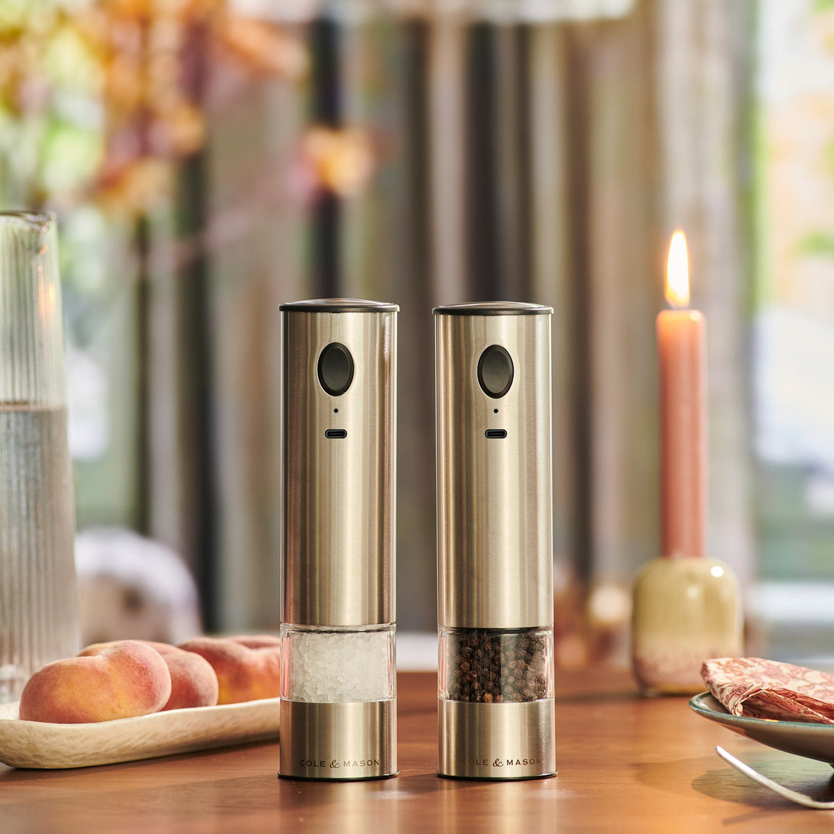 The Cole & Mason Battersea USB Electric Salt & Pepper Mills with ceramic grinders rest on a wooden table among peaches, a glass of water, a pink candle, and food. Warm lighting and a blurred background create an inviting atmosphere.