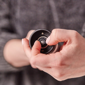 A person uses one hand to adjust the settings on the Cole & Mason Brixton Electric Mill Matt Black GS, a small, round electronic device, with their thumb and index finger. The background is out of focus.