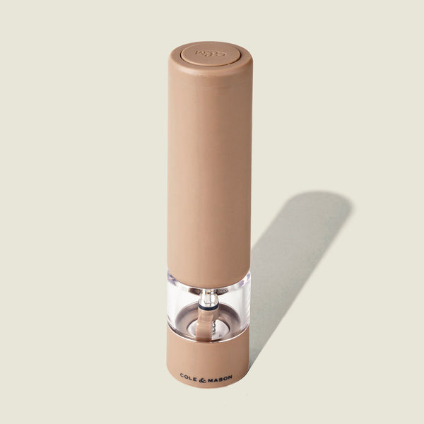 A tan, cylindrical Cole & Mason Brixton Electric Mill Stone GS pepper grinder with a transparent mid-section displaying peppercorns inside, featuring adjustable grind settings for customized seasoning, stands upright on a light background with the brand name visible at its base.