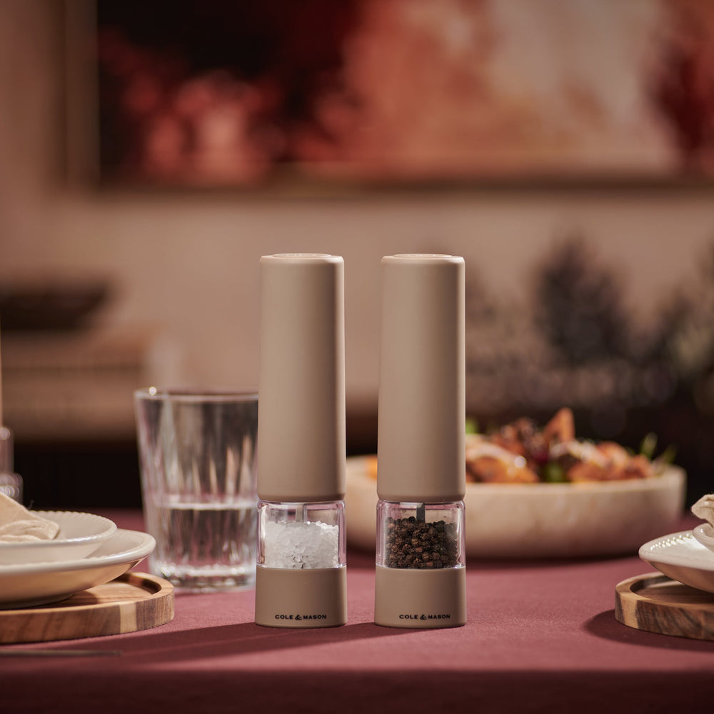 Two beige Cole & Mason Brixton Electric Mill Stone GS grinders with transparent midsections and the brand name printed at their bases, featuring adjustable grind settings and durable ceramic grinding cores, stand side by side on a light neutral background.