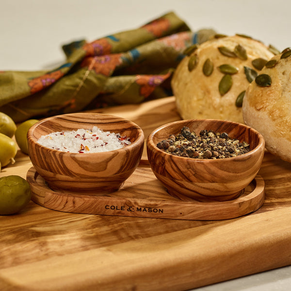 The Cole & Mason Portobello Olive Wood Pinch Pot Set features two uniquely grained pots on a matching tray, perfect for serving sea salt, pepper flakes, or mixed peppercorns alongside bread and green olives.