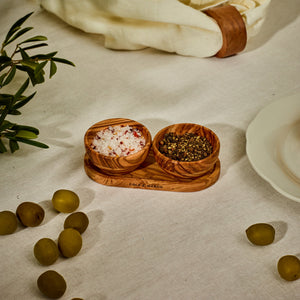 The Cole & Mason Portobello Olive Wood Pinch Pot Set features two uniquely grained pots on a seasoning tray, perfect for salt and pepper. Shown on a white cloth next to green olives, a plate, and a napkin with a leather ring.