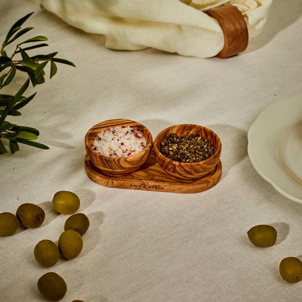 The Cole & Mason Portobello Olive Wood Pinch Pot Set features two uniquely grained pots on a seasoning tray, perfect for salt and pepper. Shown on a white cloth next to green olives, a plate, and a napkin with a leather ring.