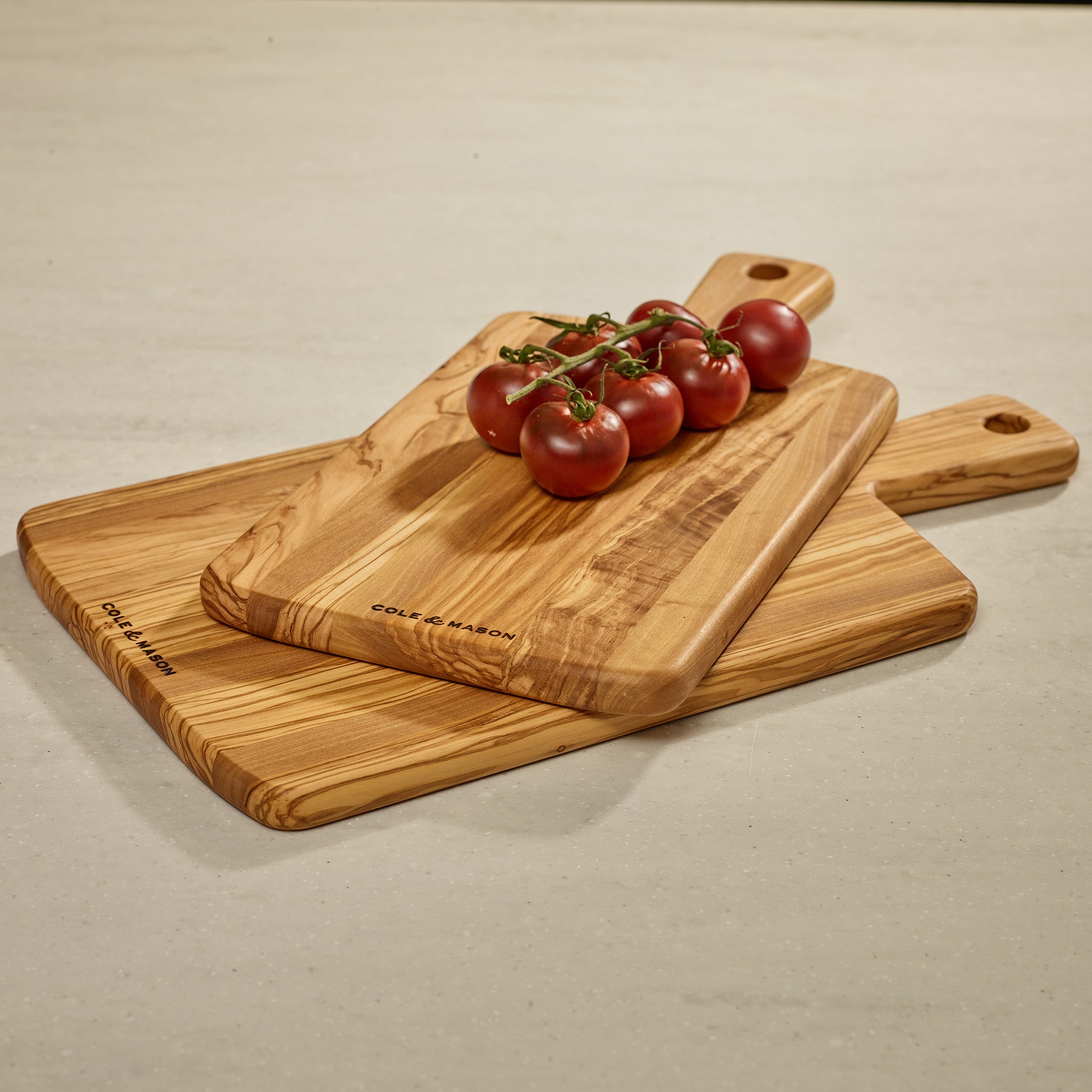 Three Cole & Mason Portobello Olive Wood Serving Boards are stacked on a countertop, topped with vine-ripened tomatoes. Each board has a handle with a hole, ideal for use as an antipasti or cheese board.