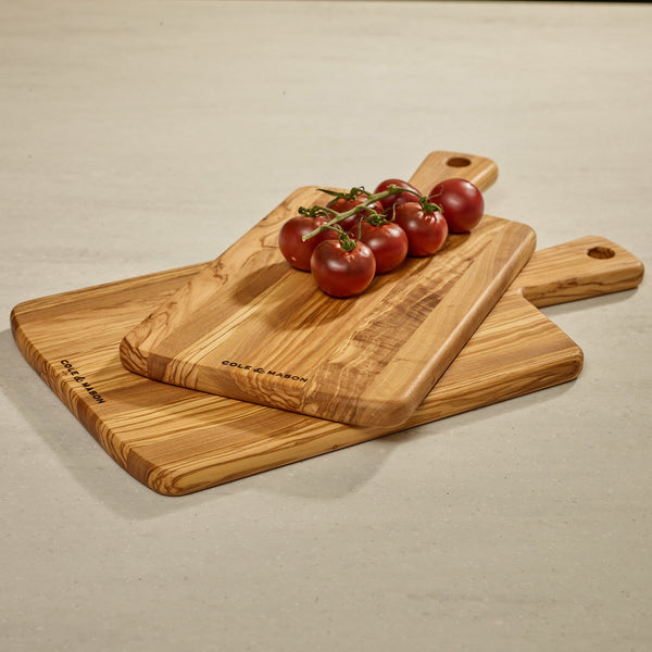 Three Cole & Mason Portobello Olive Wood Serving Boards are stacked on a countertop, topped with vine-ripened tomatoes. Each board has a handle with a hole, ideal for use as an antipasti or cheese board.