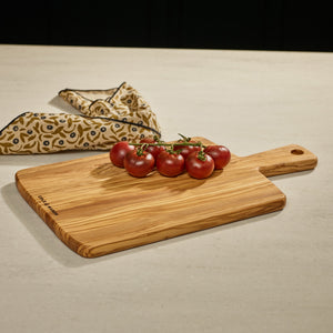 A cluster of ripe red tomatoes rests on a Cole & Mason Portobello Olive Wood Serving Board with handle, set atop a light countertop. A patterned cloth in the background adds charm reminiscent of an antipasti presentation.