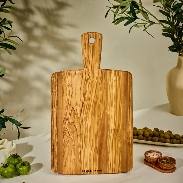The Cole & Mason Portobello Olive Wood Serving Board stands upright on a table, surrounded by green olives, a vase with leafy branches, green tomatoes, and small wooden bowls holding salt and pepper in warm natural light.