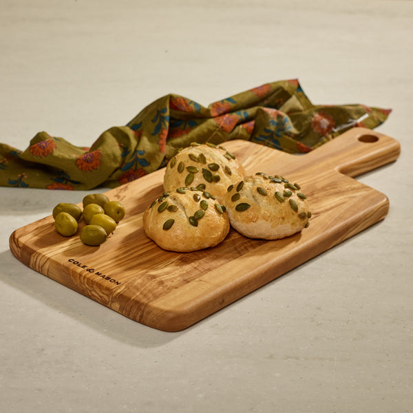 Three round bread rolls topped with pumpkin seeds and green olives are arranged on a Cole & Mason Portobello Olive Wood Serving Board, with a green floral cloth in the background on a light countertop.