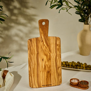 The Cole & Mason Portobello Olive Wood Serving Board stands upright on a table, accompanied by green olives, a small bowl of salt and pepper, and a vase with leafy branches in warm, natural light.