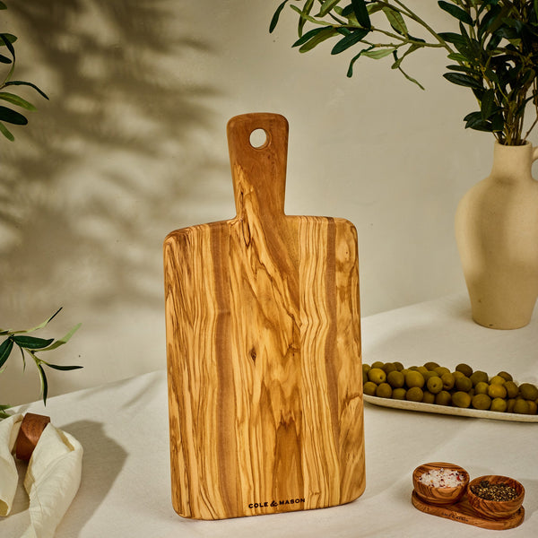 The Cole & Mason Portobello Olive Wood Serving Board stands upright on a table, accompanied by green olives, a small bowl of salt and pepper, and a vase with leafy branches in warm, natural light.
