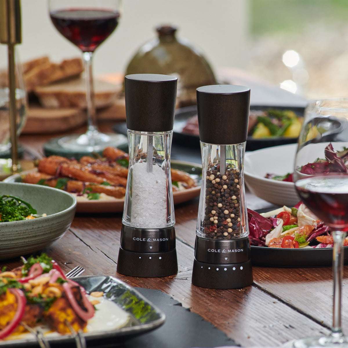 Two glass and dark wood salt and pepper grinders sit on a wooden dining table set with plates of food, wine glasses, and bread in the background.