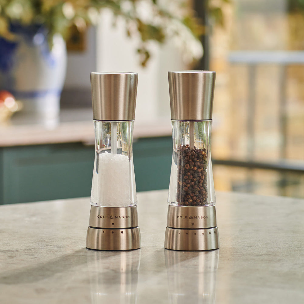 Two stainless steel and clear acrylic grinders, one filled with coarse salt and the other with whole black peppercorns, are placed side by side on a kitchen countertop.