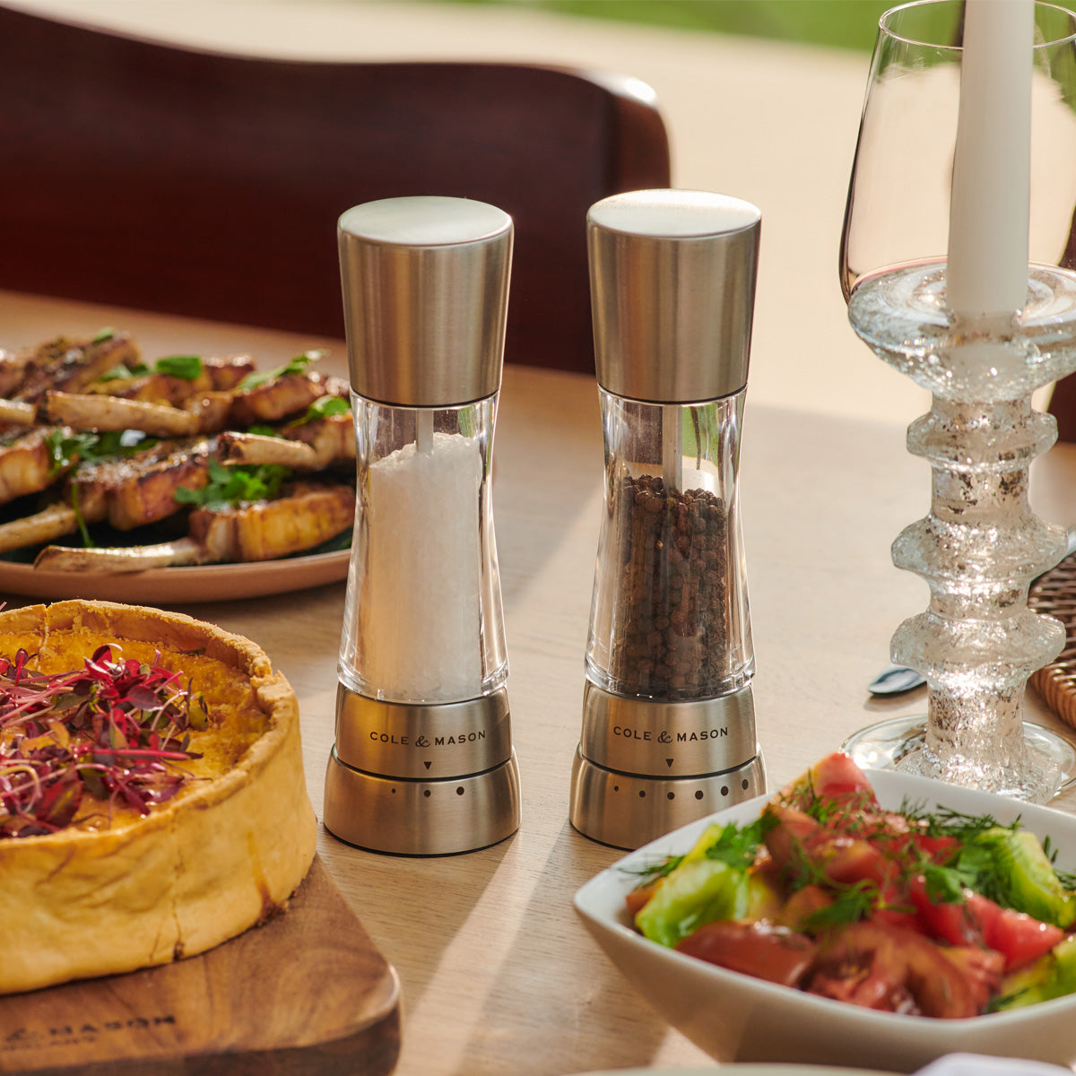A close-up of salt and pepper grinders on a dining table set with a salad, a savory pie, grilled meat, a candlestick, and a wine glass, all in warm natural light.