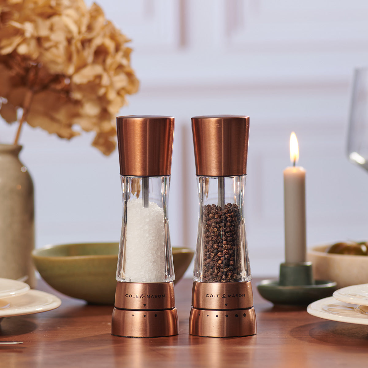 Two copper salt and pepper grinders sit on a set dining table, surrounded by dishes, a lit candle, and a vase with dried flowers in the background.