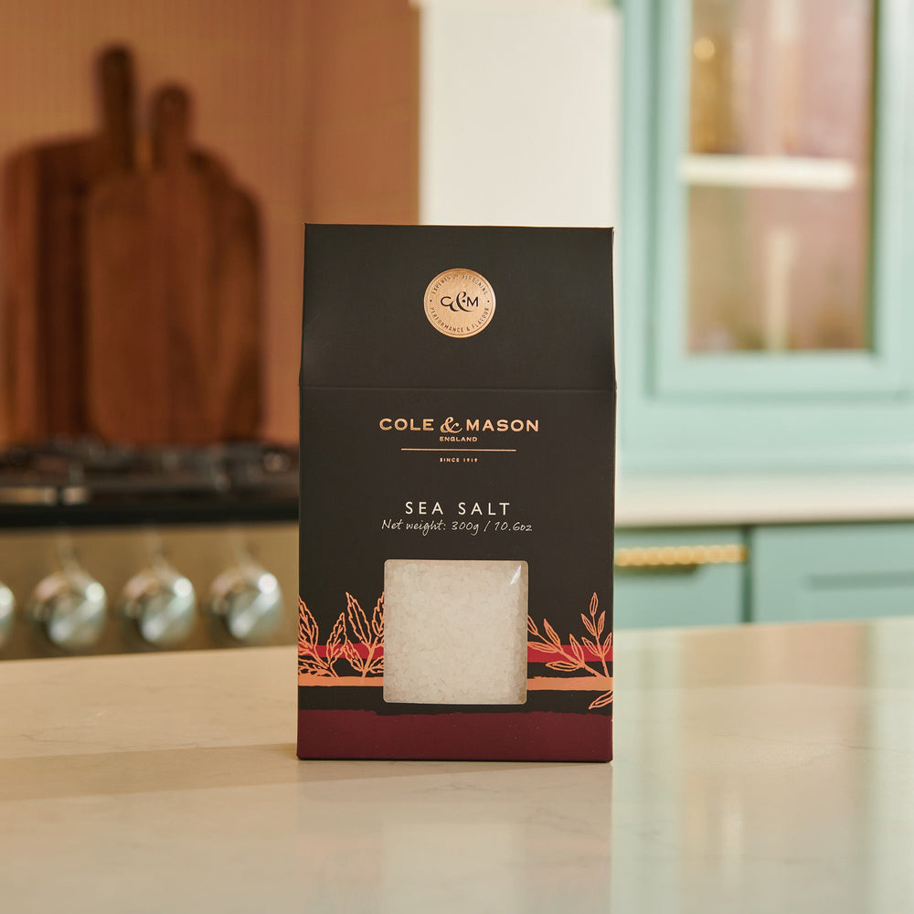 The Cole & Mason Sea Salt Box features a black and red design with a clear window revealing coarse sea salt crystals inside. Ideal for salt mills, the packaging includes product details and a nutrition facts label on the side.