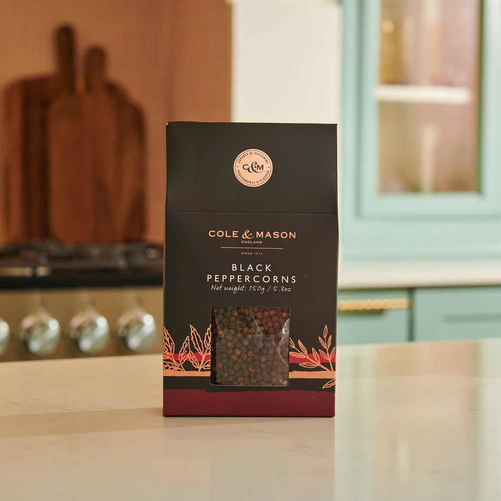The Cole & Mason Black Peppercorns Box features a rectangular black and brown design with a clear window to view the peppercorns. Ideal for pepper mills, it ensures fresh seasoning. The front displays the brand and product name.