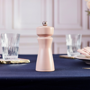 A Cole & Mason Kenton Salt & Pepper Mill in Himalayan Salt—a pale pink hue reminiscent of Himalayan salt—sits on a navy blue tablecloth, accompanied by two clear drinking glasses and plates, against a white paneled wall; its durable ceramic grinder ensures lasting performance.