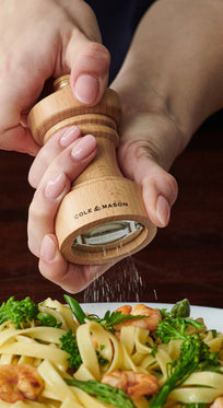 A person grinds fresh pepper onto a plate of pasta with shrimp and broccoli using a wooden pepper mill labeled Cole & Mason.