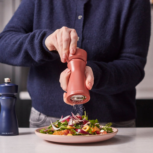 A person seasons a colorful salad on a plate using the Cole & Mason Kenton Salt & Pepper Mill in Cinnamon, which features a ceramic grinder. Another Kenton Salt & Pepper Mill rests on the counter nearby, complementing the kitchen accessories with its stylish design. The person is dressed in a dark sweater and light-colored pants.
