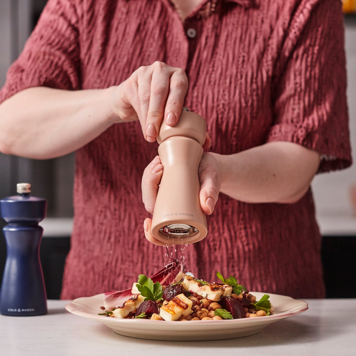 A person in a red shirt grinds pepper onto a plate of food using the Cole & Mason Kenton Salt & Pepper Mill in Himalayan Salt, featuring a ceramic grinder for an elegant touch. Meanwhile, another Kenton mill in dark blue—ideal for Himalayan pink salt—rests on the counter nearby.