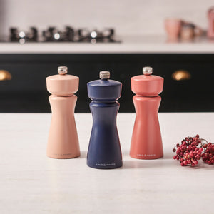 A Cole & Mason Kenton Salt & Pepper Mill in Persian Blue Salt stands on a kitchen counter alongside two other mills in peach and coral shades, all featuring ceramic grinders. Pink peppercorns are scattered nearby, and a stove is visible in the background.