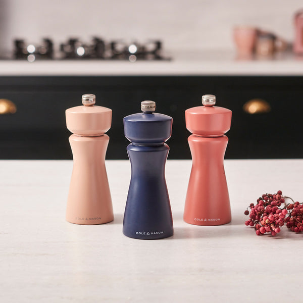 A Cole & Mason Kenton Salt & Pepper Mill in Persian Blue Salt stands on a kitchen counter alongside two other mills in peach and coral shades, all featuring ceramic grinders. Pink peppercorns are scattered nearby, and a stove is visible in the background.