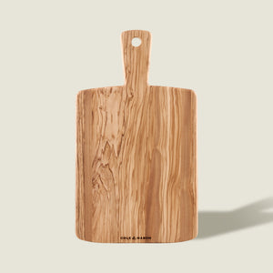 The Cole & Mason Portobello Olive Wood Serving Board, with a handle and hanging hole, showcases an elegant natural grain—ideal as a stylish antipasti board and shown upright on a light background.