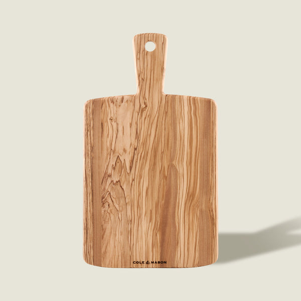 The Cole & Mason Portobello Olive Wood Serving Board, with a handle and hanging hole, showcases an elegant natural grain—ideal as a stylish antipasti board and shown upright on a light background.