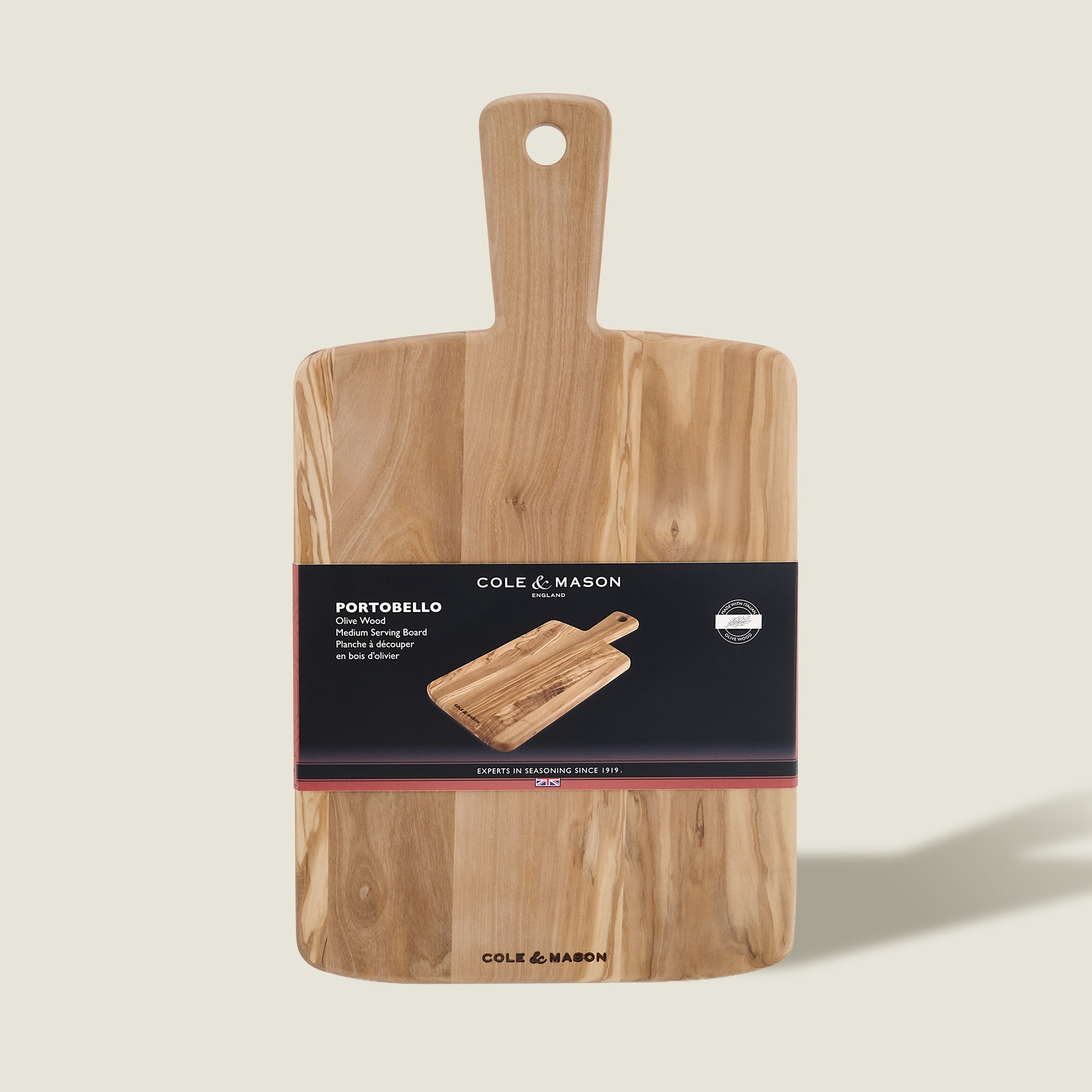 A rectangular olive wood serving board with a handle, made by Cole & Mason. Labeled "Portobello Olive Wood Serving Boards," it features a black band with product info and an image of the antipasti board.