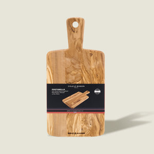 A rectangular Cole & Mason Portobello Olive Wood Serving Board with a handle stands upright against a light background, showcasing its natural, varied wood grain and rustic charm.