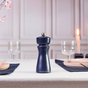 A Kenton Salt & Pepper Mill in Persian Blue Salt by Cole & Mason, featuring a ceramic mill and adjustable grind settings for perfect seasoning, sits on a set dining table adorned with glassware, plates, a lit candle, and folded napkins. White paneled walls and soft lighting create an inviting backdrop.