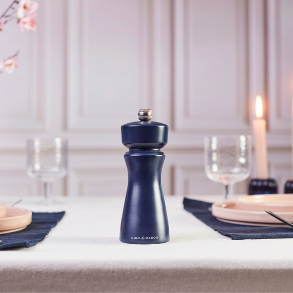 A Kenton Salt & Pepper Mill in Persian Blue Salt by Cole & Mason, featuring a ceramic mill and adjustable grind settings for perfect seasoning, sits on a set dining table adorned with glassware, plates, a lit candle, and folded napkins. White paneled walls and soft lighting create an inviting backdrop.