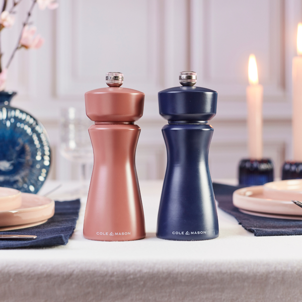 Two Cole & Mason Kenton Salt & Pepper Mills in Persian Blue Salt, featuring a ceramic mill and adjustable grind settings, are placed side by side on a set dining table with candles and plates in the background.