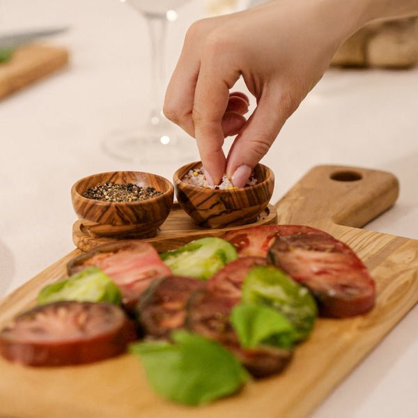 A hand sprinkles seasoning from the Cole & Mason Portobello Olive Wood Pinch Pot Set onto sliced heirloom tomatoes and basil on a cutting board, displaying the set’s unique grain patterns. A seasoning tray with a pepper bowl is nearby.