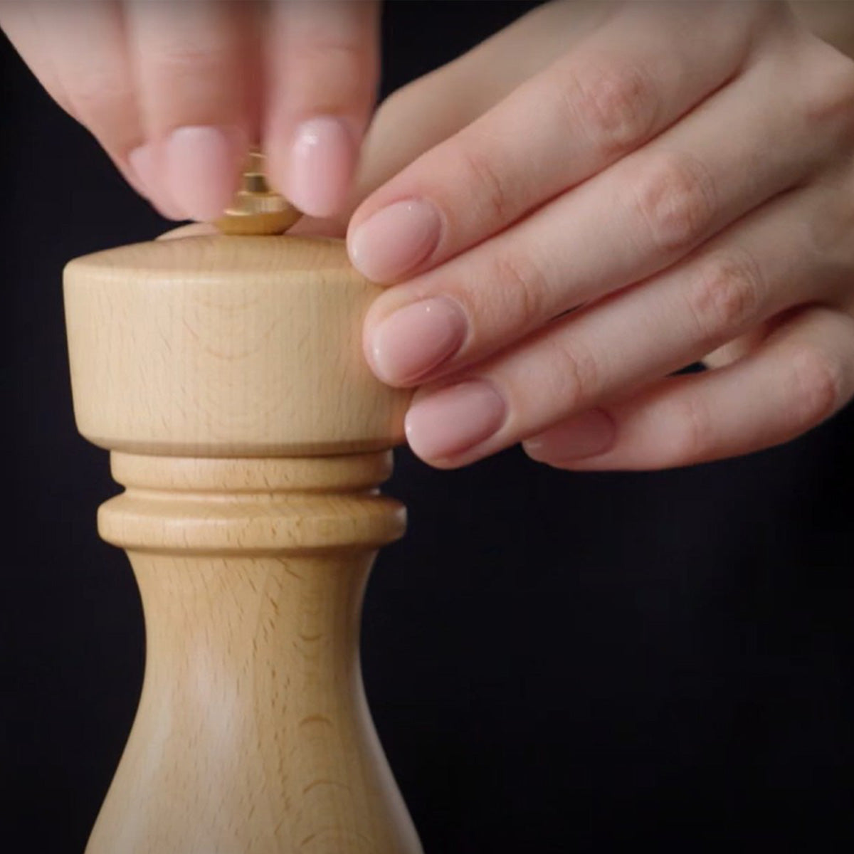 Close-up of hands with neatly manicured nails twisting the top of a wooden pepper grinder against a dark background.