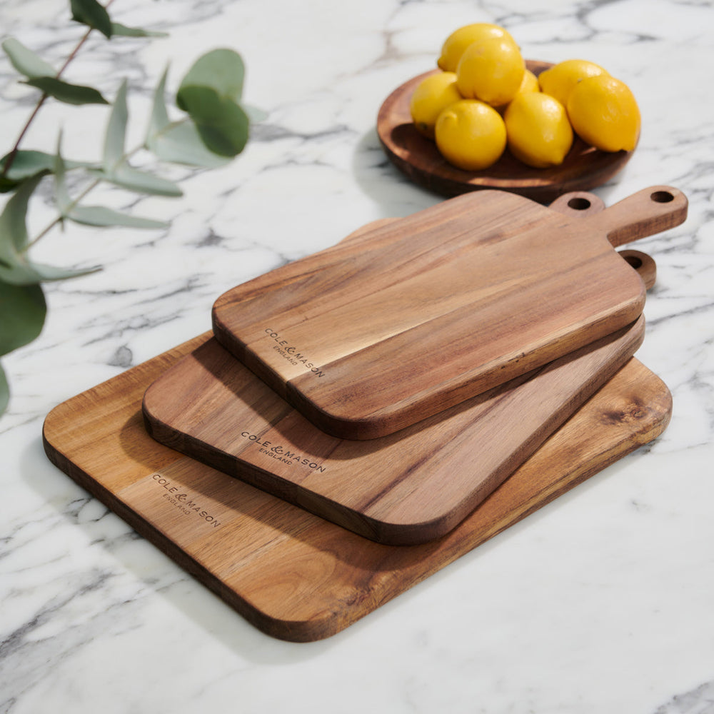 Three wooden cutting boards of varying sizes are stacked on a marble countertop. In the background, a wooden plate holds several yellow lemons, and a sprig of green leaves is visible on the left.