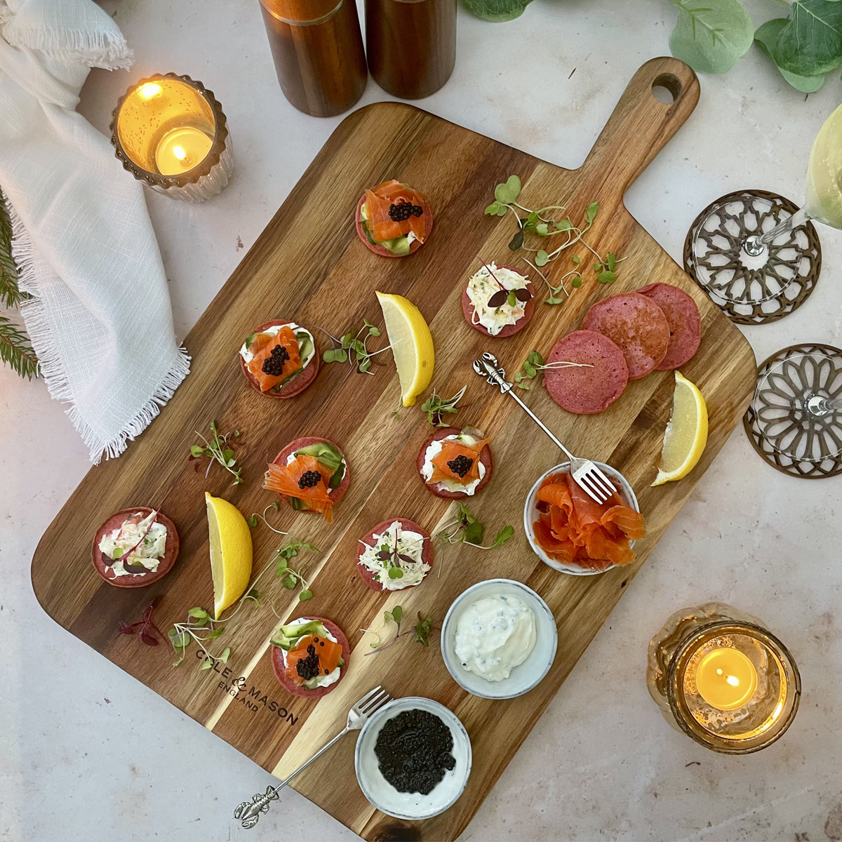 The Cole & Mason 2 Piece Barkway Board Bundle displays appetizers, lemon wedges, smoked salmon, salami, dips, caviar, and microgreens. Surrounded by candles on marble with a white cloth, it adds elegance to any gathering.