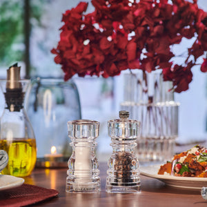 A modern table setting displays the Cole & Mason London Acrylic Salt Shaker & Pepper Mill Set on a wooden table beside a plate of food, a bottle of olive oil, a lit candle, and a vase with red flowers in the background.