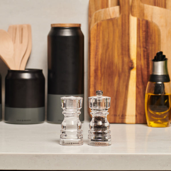 The Cole & Mason London Acrylic Salt Shaker & Pepper Mill Set adds a contemporary touch to your kitchen countertop among utensils, cutting boards, and a bottle of olive oil.