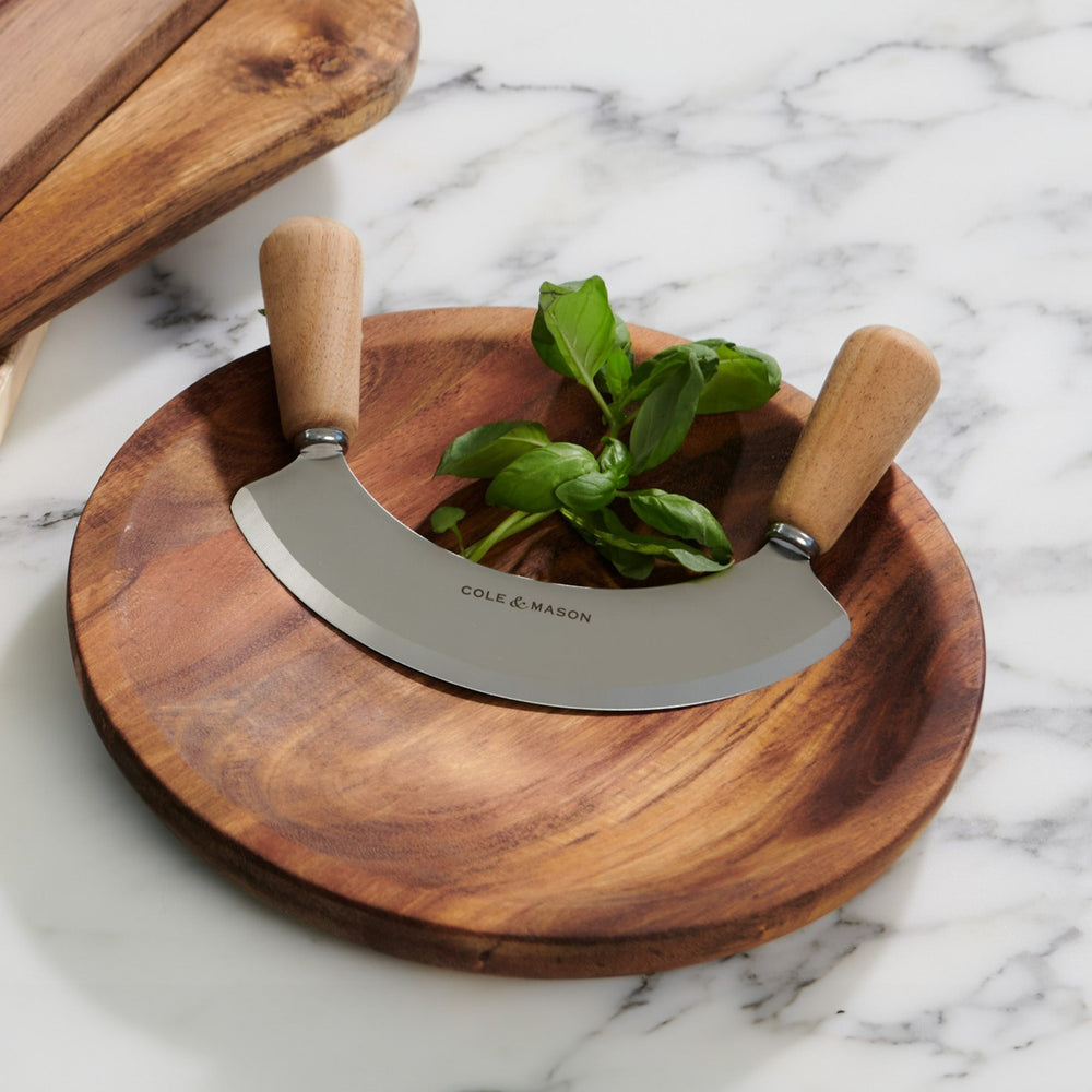 The Cole & Mason Ashden Mezzaluna and Board includes a wooden chopping board with a curved stainless steel mezzaluna, featuring wooden handles—ideal for efficient food preparation.