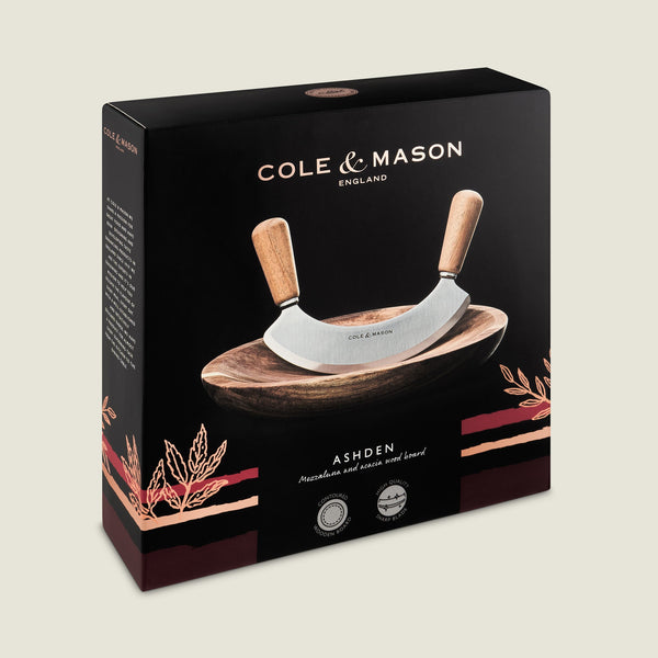 The Cole & Mason Ashden Mezzaluna and Board set features a curved chopping tool with wooden handles on an acacia wood board, displayed in stylish packaging with gold and red accents. Ideal for quick, efficient food prep.