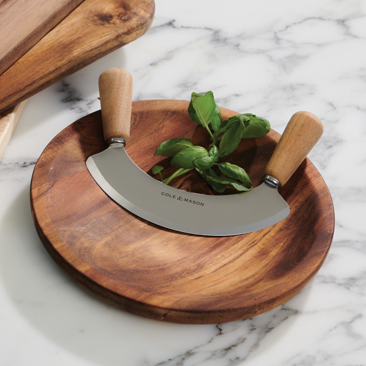 A mezzaluna herb chopper with wooden handles rests on a round wooden cutting board, with fresh basil leaves placed underneath, all on a white marble countertop.
