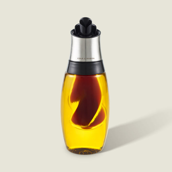 The Cole & Mason Bristol Duo Oil & Vinegar Pourer features clear glass with a metal top and black spout for mess-free dispensing. Filled with golden oil and a red chili pepper, it casts a soft shadow on a light background.