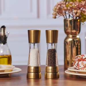 Two Cole & Mason Derwent Gold Salt & Pepper Mills with advanced grinding technology sit on a wooden dining table set for dining, adding elegance alongside plates, an oil dispenser, and a gold vase of flowers.