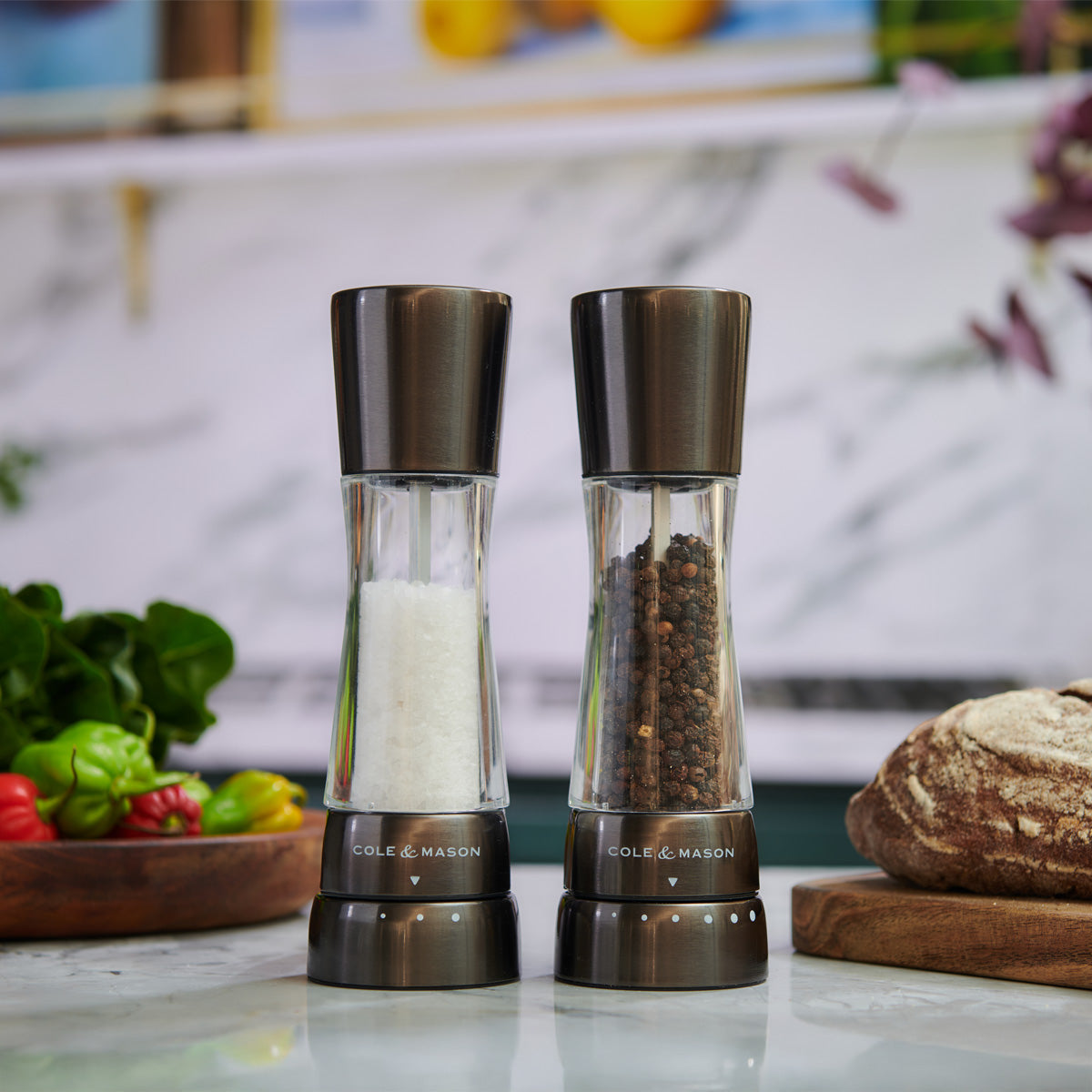 Two Cole & Mason grinders filled with salt and black pepper sit side by side on a kitchen counter, surrounded by fresh vegetables and a loaf of bread.