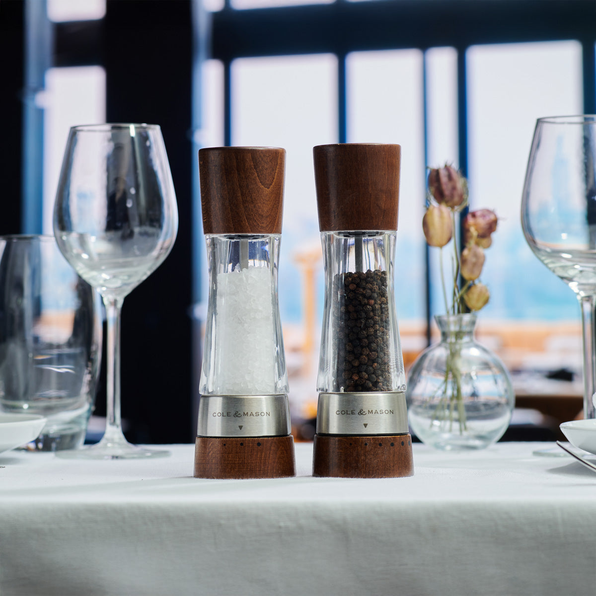 A salt grinder and a pepper grinder sit on a white tablecloth, surrounded by empty wine glasses and a small vase with dried flowers, with large windows and a blurred outdoor view in the background.