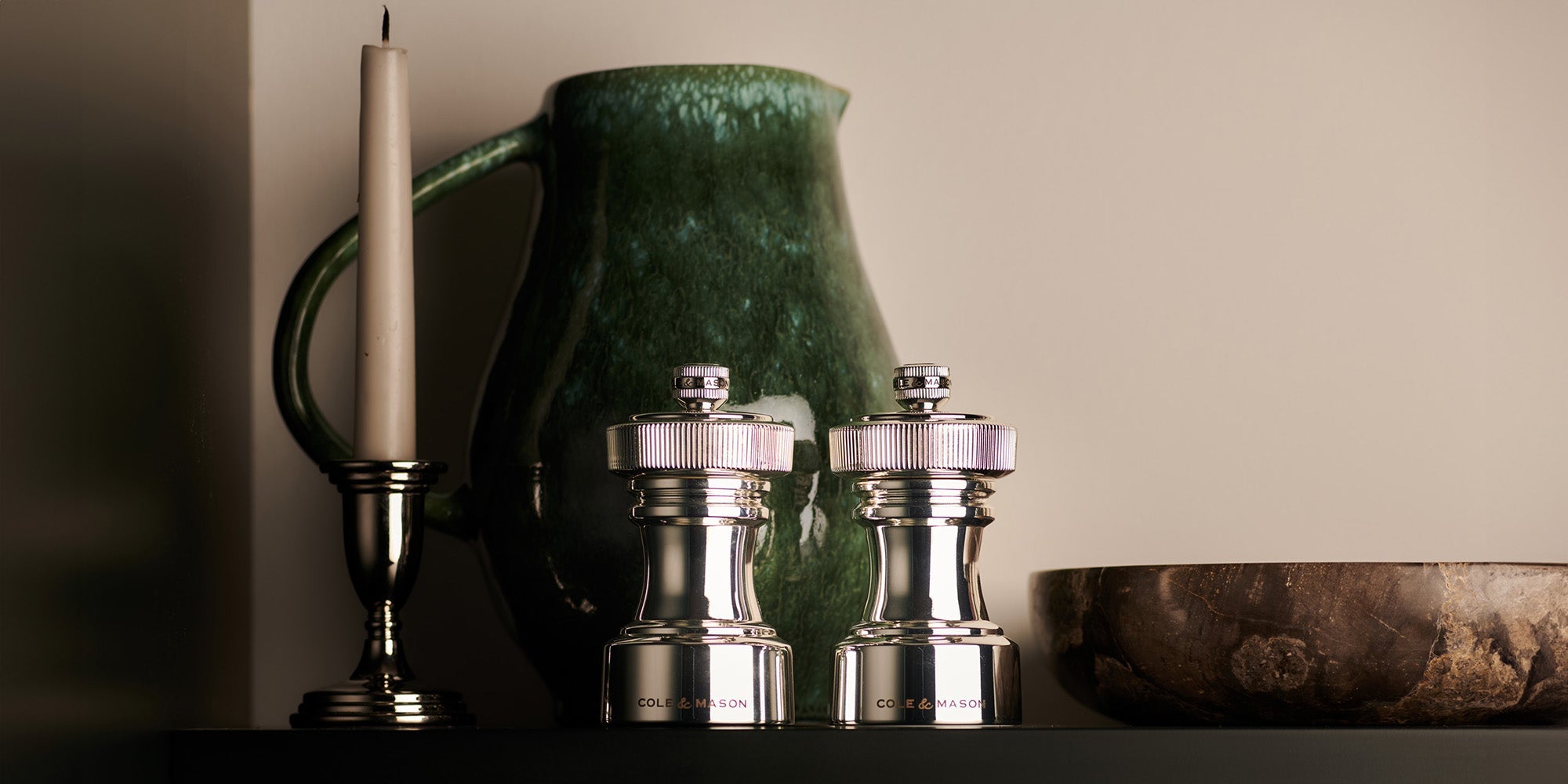 A silver candlestick with a white candle, a green ceramic jug, two metal salt and pepper grinders, and a brown bowl are displayed on a dark shelf against a beige wall.