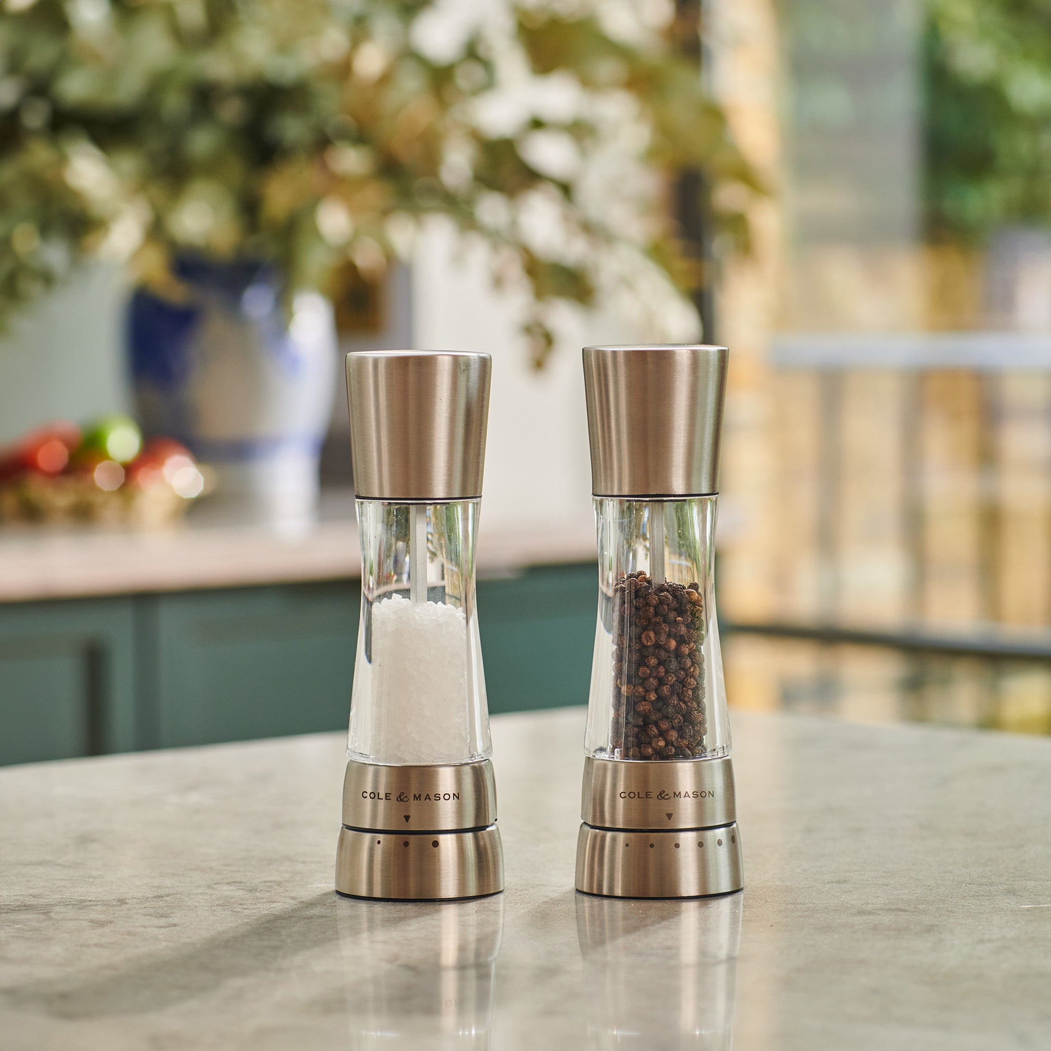 Two clear and stainless steel mills, one filled with coarse salt and the other with whole black peppercorns, sit side by side on a kitchen countertop with a blurred background.