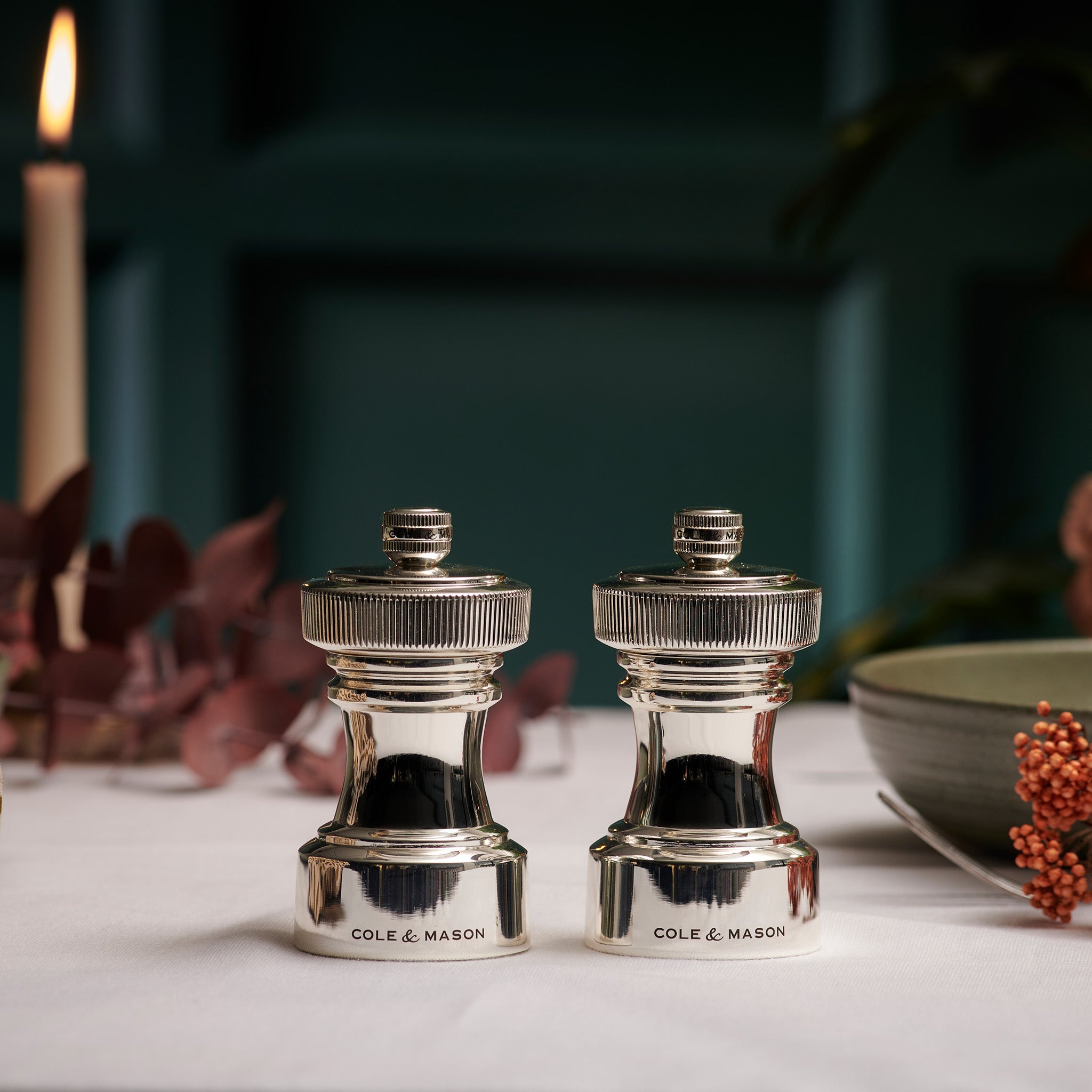 Two shiny silver Cole & Mason salt and pepper grinders sit side by side on a white tablecloth, with a lit candle, green bowl, and autumnal decor in the background.