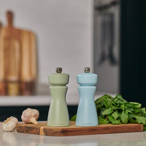 The Cole & Mason Kenton Blue Salt & Pepper Mill, with a refillable design and durable ceramic mechanism, sits on a wooden board alongside fresh mint and garlic, adding a pop of color to the kitchen counter.